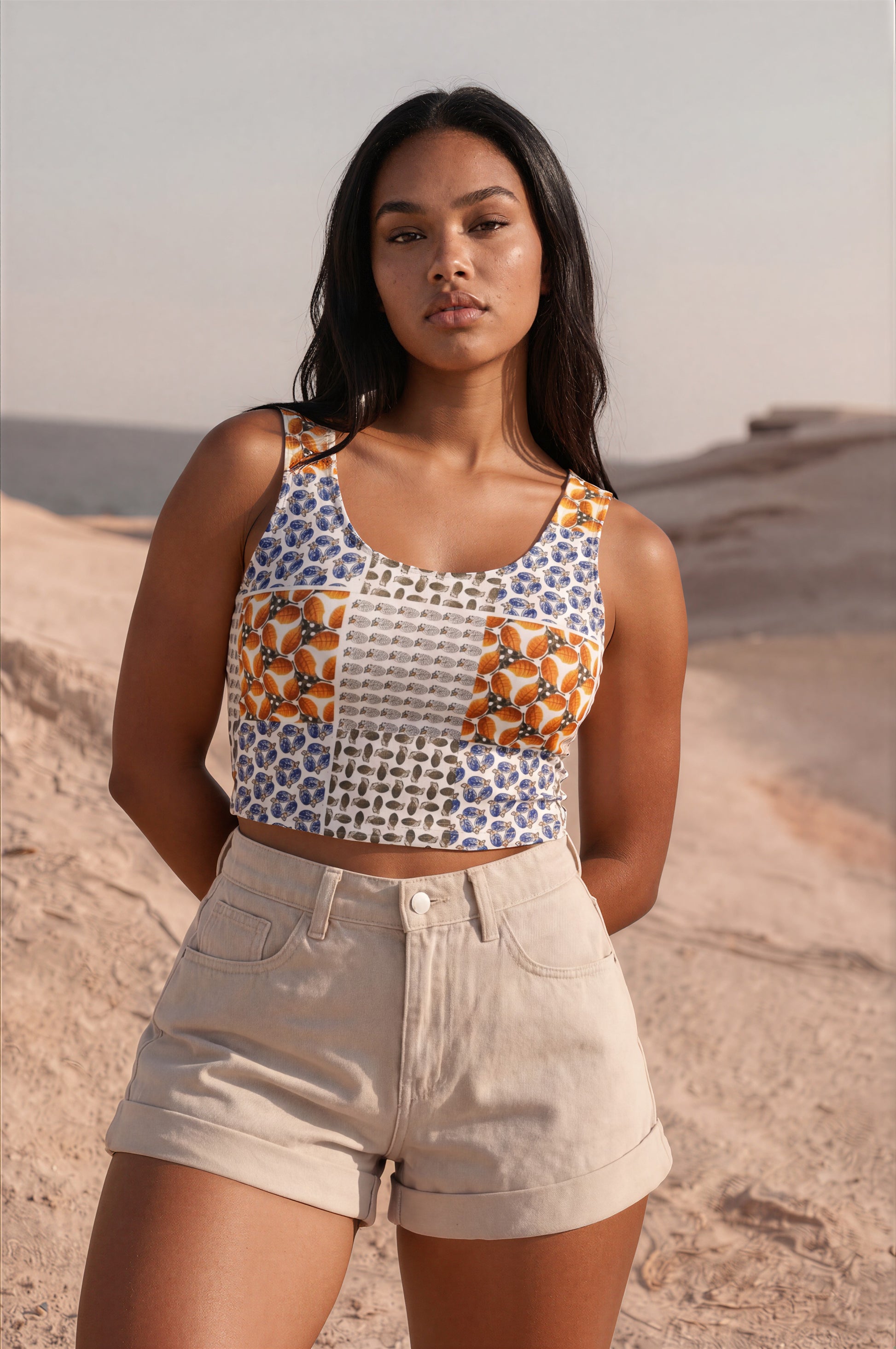 A woman standing outdoors in a desert landscape wearing a sleeveless patchwork crop top with blue, orange, and white patterns, paired with light beige shorts.