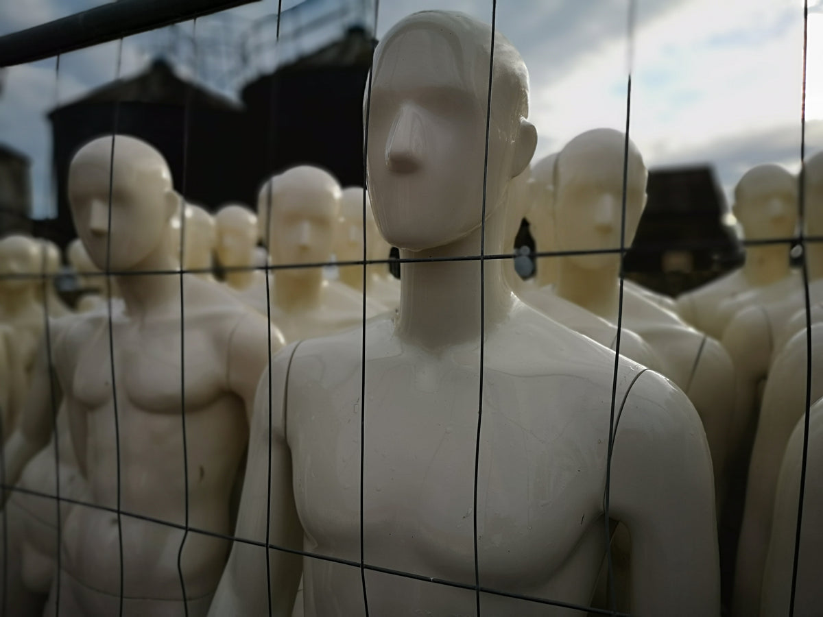 A row of white male mannequins standing behind a black wire mesh fence, photographed outdoors with a cloudy sky and dark buildings in the background.