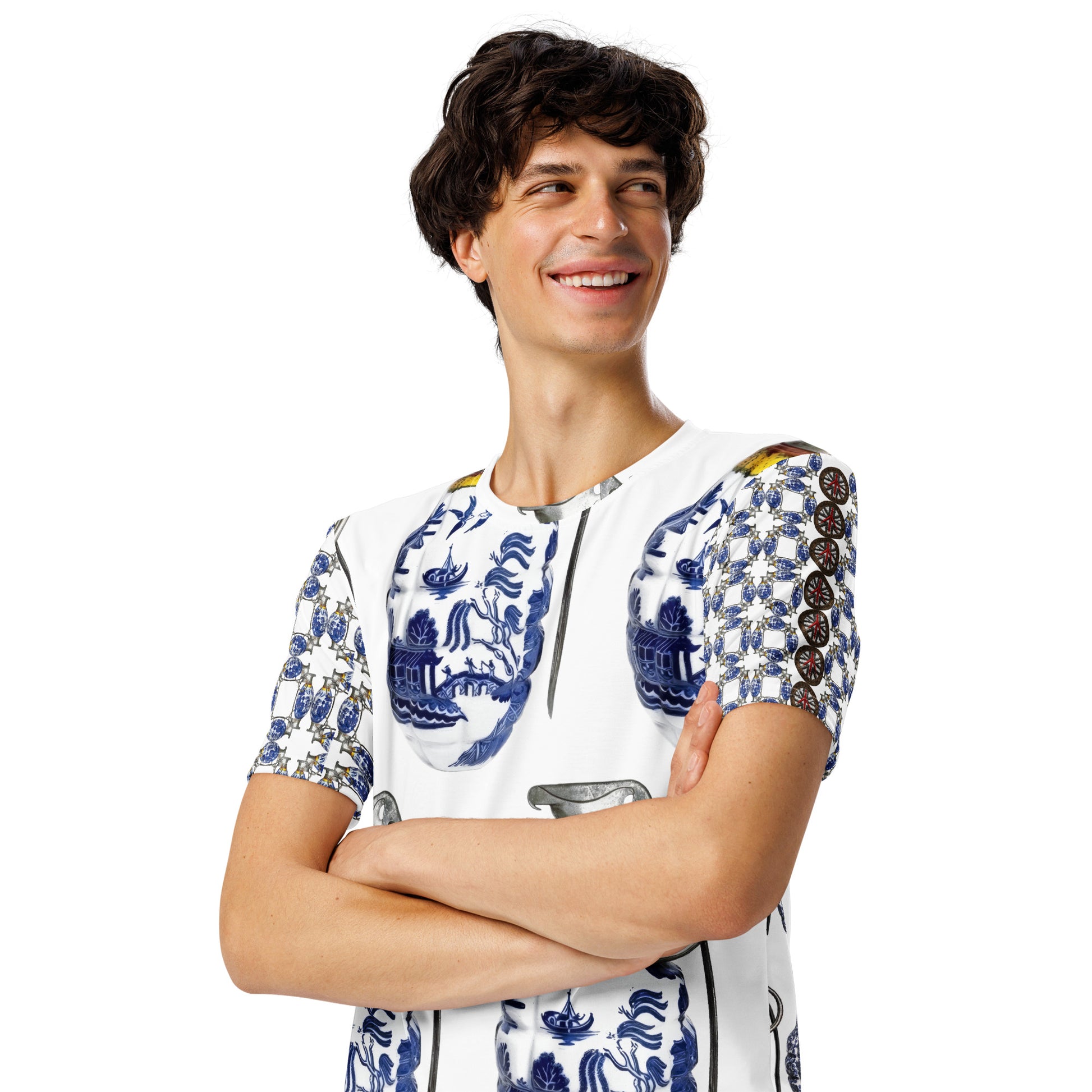 A smiling man with short dark curly hair wearing a white short-sleeve t-shirt with a blue and white grenade pattern, standing with arms crossed against a plain white background.