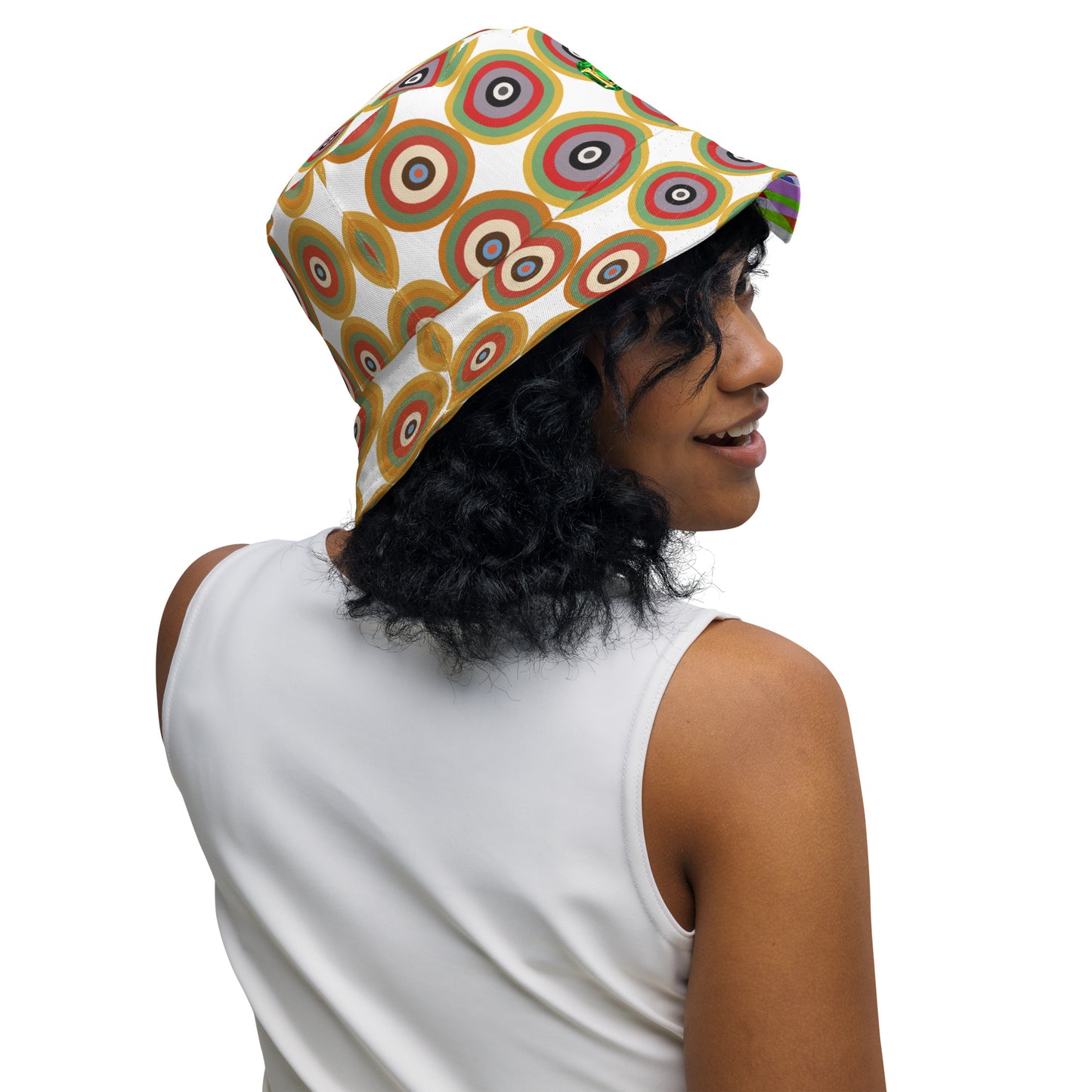 Woman wearing a  bucket hat featuring a retro circular target pattern on the crown in shades of green, red, brown, and beige on a white background, with a colorful geometric pattern on the inside. Reversible and breathable.
