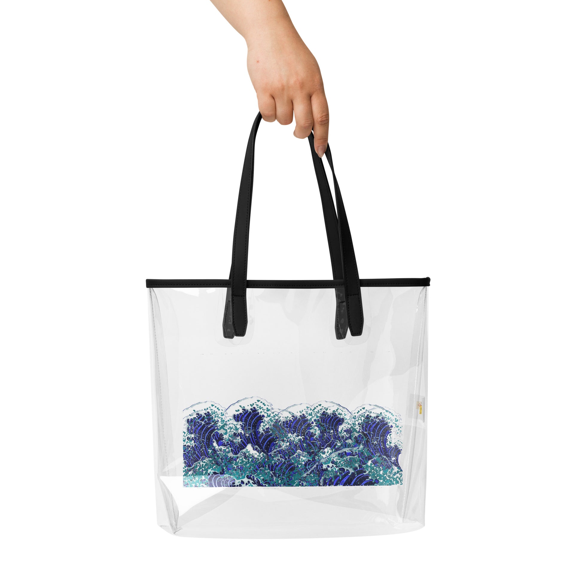 A clear plastic beach tote bag with black handles and a blue and white wave graphic near the bottom, held by a hand against a white background.