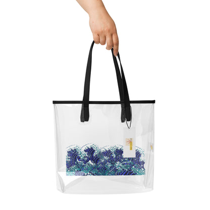 A hand holding a clear plastic tote bag with black handles and a blue and white wave print on the front, shown against a plain white background.
