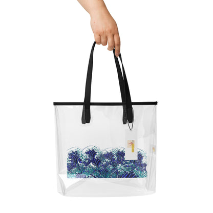 A hand holding a clear plastic tote bag with black handles and a blue and white wave print on the front, shown against a plain white background.